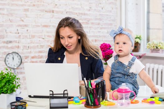 Working together is so fun Cheerful young beautiful businesswoman looking at laptop while sitting at her working place with her little daughter