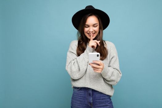 Attractive young smiling woman wearing black hat and grey sweater holding smartphone looking down showing shhh gesture isolated on background.Copy space