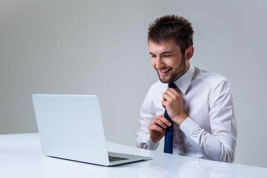 young man smiles and straightens his tie using laptop computer sitting at the table. Office clothing