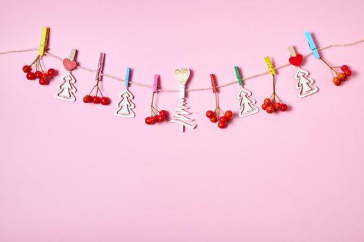 Figurines of wooden Christmas trees and rowan berries are hung with clothespins on a rope on a pink background
