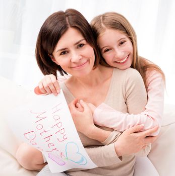 Lovely little girl hugging her mother and looking at camera. Girl holding creative handmade greeting card for mother. Daughter congratulates mom and gives her postcard for Happy Mother's day
