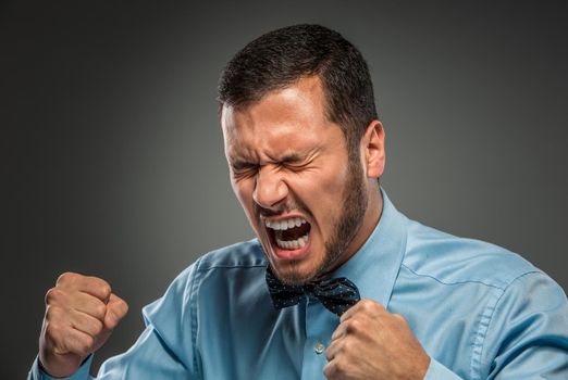 Portrait of angry upset young man in blue shirt and butterfly tie with fists up yelling isolated on gray studio background. Negative human emotion, facial expression. Closeup