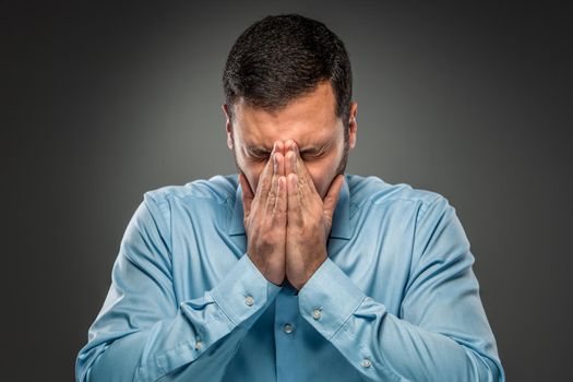 Closeup portrait of upset man in blue shirt and butterfly tie covering his face by hands, isolated on gray background. Feeling sick and tired - Human emotion.