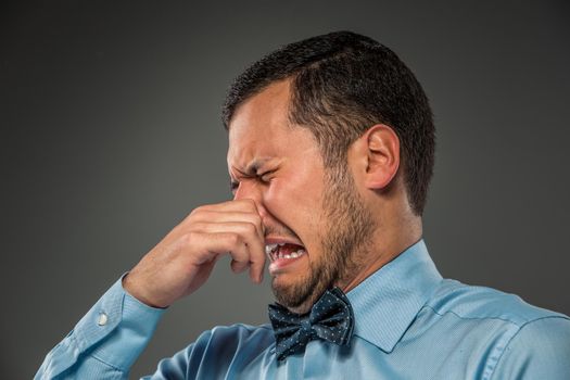 Young man in blue shirt and butterfly tie, with disgusted expression closing his nostrils with his fingers, isolated on gray background. Closeup. Mimicry. Gesture.