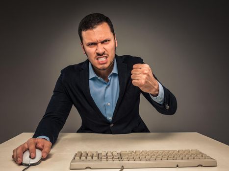 Portrait of angry upset young man in blue shirt and jacket with fists up yelling sitting at a desk near a computer isolated on gray background. Negative human emotion, facial expression. Closeup