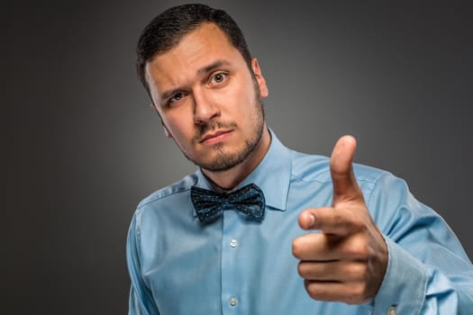 Closeup portrait of man in blue shirt and butterfly tie is gesturing with his hand, pointing finger at camera isolated on gray background.