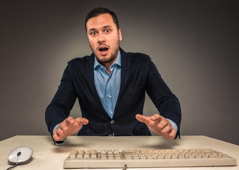 Portrait young man in blue shirt and jacket looking at the camera with amazement, sitting at a desk near a computer isolated on gray studio background. Human emotion, facial expression. Closeup