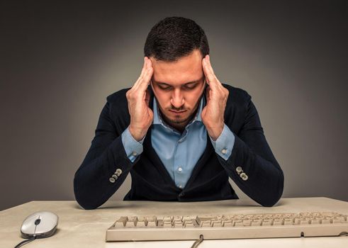 Portrait frustrated young man, hands touch the head, sitting at a desk near a computer, isolated on gray background. Feeling sick and tired - Human emotion.