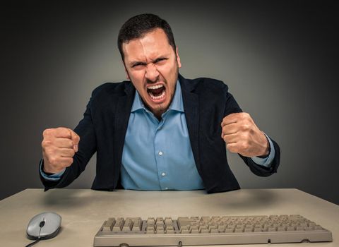 Portrait of angry upset young man in blue shirt and jacket with fists up yelling sitting at a desk near a computer isolated on gray background. Negative human emotion, facial expression. Closeup
