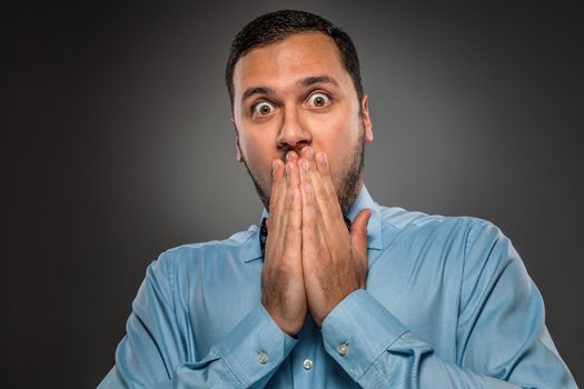 Portrait young man in blue shirt and butterfly tie looking at the camera with amazement, her hands covered her mouth isolated on gray studio background. Human emotion, facial expression. Closeup
