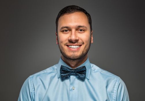 Portrait of happy smiling young man in blue shirt and butterfly tie isolated over gray studio background. Closeup