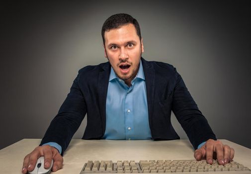 Portrait young man in blue shirt and jacket looking at the camera with amazement, sitting at a desk near a computer isolated on gray studio background. Human emotion, facial expression. Closeup