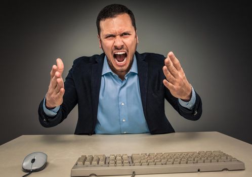 Portrait of angry upset young man in blue shirt and jacket with hands up yelling sitting at a desk near a computer isolated on gray background. Negative human emotion, facial expression. Closeup