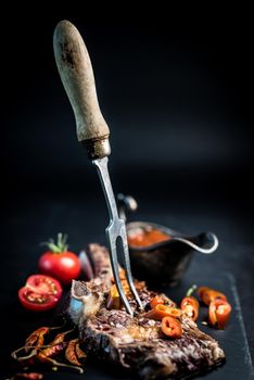 Tasty homemade beef grilled steak served with meat fork, tomatoes, chili pepper and pot of sauce on dark cooking board