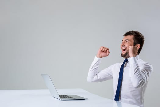 the young man behind the laptop. It uses a computer while sitting at a table. enjoys hands up. Office clothing