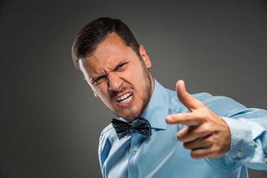 Portrait of angry upset young man in blue shirt and butterfly tie, pointing finger at camera, isolated on gray background. Negative human emotion, facial expression. Closeup