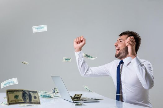 the young man behind the laptop. It uses a computer while sitting at a table. enjoys hands up. Office clothing. the top falling money, dollars