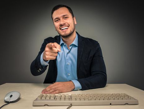 Closeup portrait of excited, energetic, happy, smiling man in blue shirt and jacket is gesturing with his hand, pointing finger at camera sitting at a desk near a computer isolated on gray background. Positive human emotion facial expression