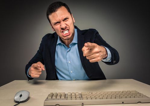 Portrait of angry upset young man in blue shirt and jacket with hands up yelling sitting at a desk near a computer isolated on gray background. Negative human emotion, facial expression. Closeup