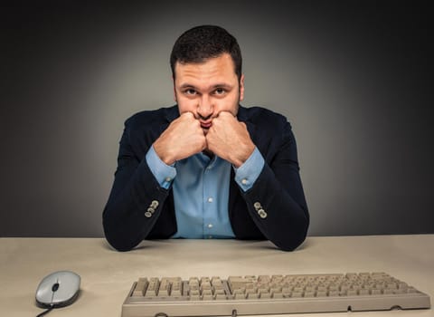 Portrait young man in blue shirt and jacket looking at the camera, hands holding his head, sitting at a desk near a computer isolated on gray studio background. Human emotion, facial expression. Closeup