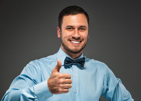 Happy smiling young man in blue shirt and butterfly tie making the ok thumbs up hand sign, gesture, isolated over gray studio background.