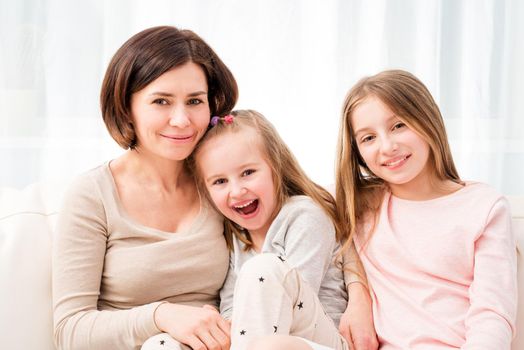 Mom embracing her cute cheerful little daughters while sitting on the sofa at home. Happy family portrait