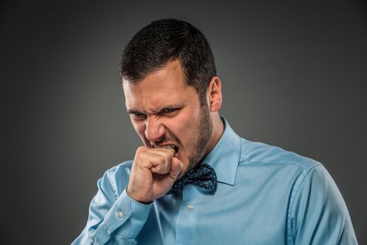 Closeup portrait of an angry guy in blue shirt and butterfly tie, biting his fist, isolated on gray background. Human emotion.