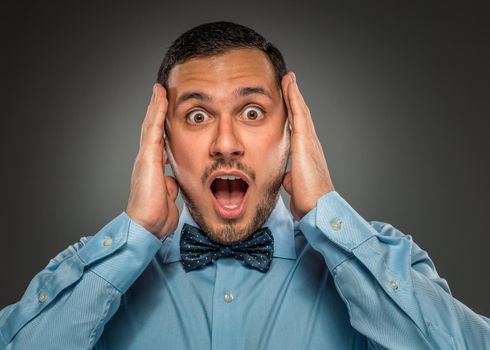 Portrait young man in blue shirt and butterfly tie looking at the camera with amazement, hands touch the head isolated on gray studio background. Human emotion, facial expression. Closeup