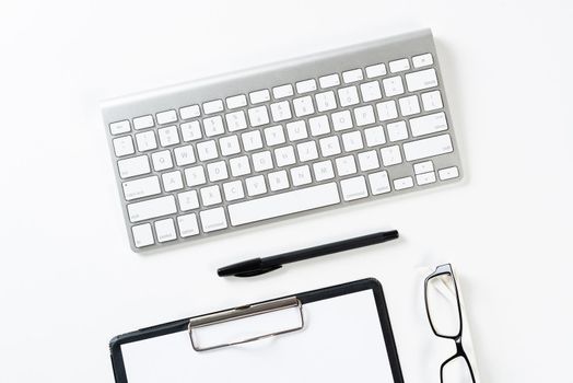 Top view of well organized workplace with office accessories. Clipboard with blank paper page, glasses and wireless keyboard on white surface. Education, creativity and working concept with copy space