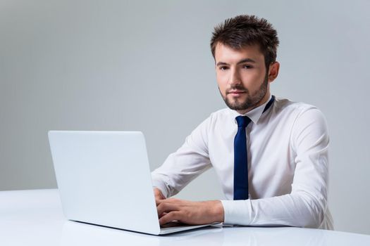 young man smiling at the camera uses a laptop computer while sitting at a table. Office clothing