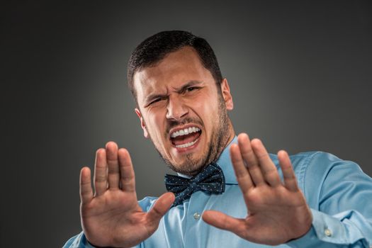 Portrait of angry upset young man in blue shirt and butterfly tie with hands up yelling isolated on gray background. Negative human emotion, hands show Stop, facial expression. Closeup