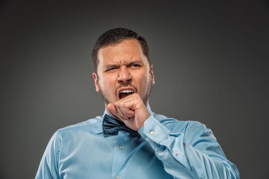 Young man in blue shirt and butterfly tie, yawns, covering her mouth isolated on gray background. Closeup