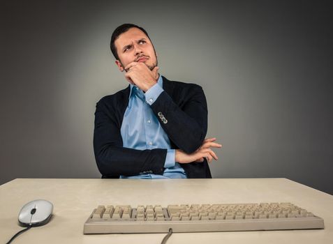 Handsome man in blue shirt and jacket looking up, sitting at a desk near a computer, isolated on gray background. Concept of the idea or warning