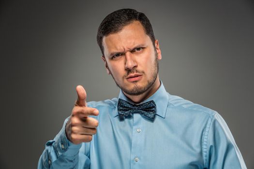 Portrait of angry upset young man in blue shirt and butterfly tie with pointing finger at camera, isolated on gray background. Negative human emotion, facial expression. Closeup
