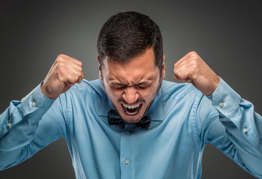 Portrait of angry upset young man in blue shirt and butterfly tie with fists up yelling isolated on gray studio background. Negative human emotion, facial expression. Closeup