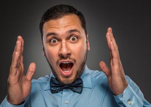 Portrait young man in blue shirt and butterfly tie looking at the camera with amazement, isolated on gray studio background. Human emotion, facial expression. Closeup