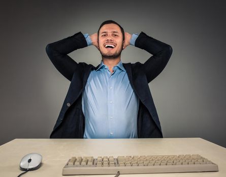 Closeup portrait of excited, energetic, happy, man in blue shirt and butterfly tie winning, his hands kept his head sitting at a desk near a computer isolated on gray background. Positive human emotion facial expression