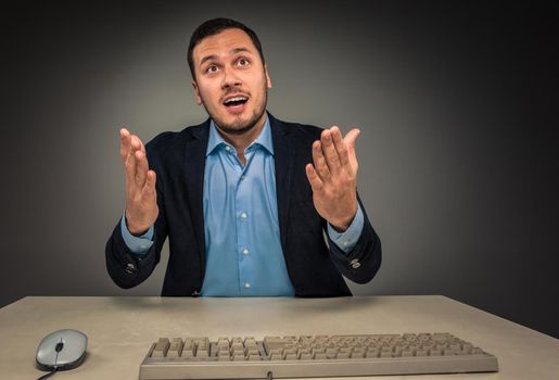 Portrait young man in blue shirt and jacket looking at the camera, hands raised, sitting at a desk near a computer isolated on gray studio background. Human emotion, facial expression. Closeup