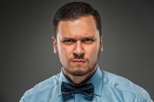 Portrait young man in blue shirt and butterfly tie looking at the camera with suspicion, isolated on gray studio background. Negative human emotion, facial expression. Closeup