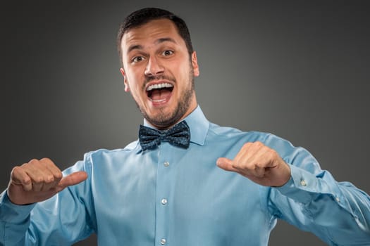 Closeup portrait of excited, energetic, happy, smiling man in blue shirt and butterfly tie is gesturing with his hand, pointing finger shows itself isolated on gray background. Positive human emotion facial expression