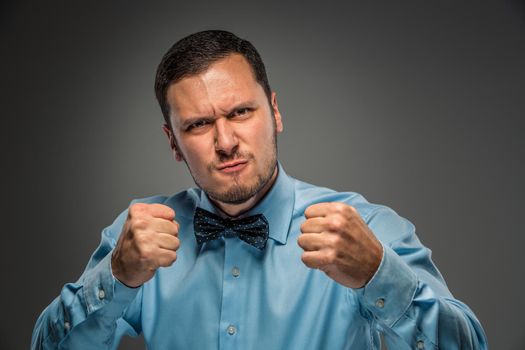 Portrait of angry upset young man in blue shirt and butterfly tie with fists up isolated on gray studio background. Negative human emotion, facial expression. Closeup