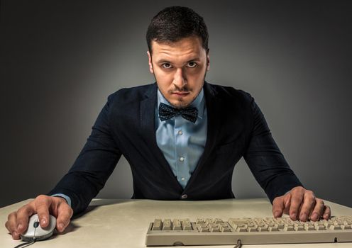 Portrait young man in blue shirt and jacket looking at the camera, sitting at a desk near a computer isolated on gray studio background. Human emotion, facial expression. Closeup