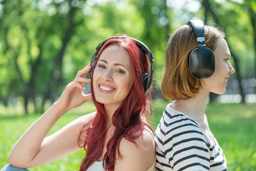 Two young women listen to music in the park. Sitting back to back