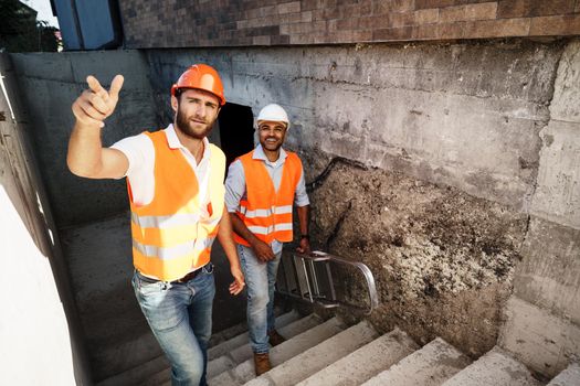 Two young male engineers in uniform and hardhats working at construction site, close up