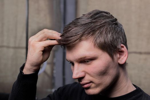 A young guy portrait of Caucasian appearance touches with his hand, his long dark hair necessary for a haircut and in need of hairdressing services in a barbershop.