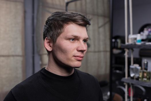 A young guy portrait of Caucasian appearance with dark hair in a black sweatshirt against the background of a hairdresser.