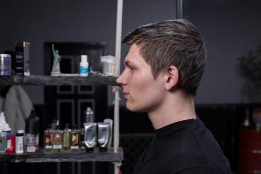 Portrait in profile of a young guy of Caucasian appearance with dark hair in a black sweatshirt against the background of a hairdresser. The need for hairdressing services and cutting overgrown hair.