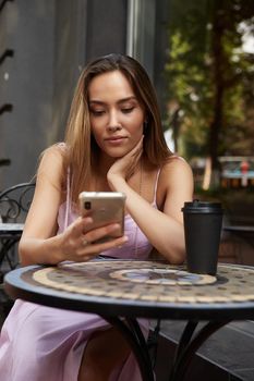 young well dressed woman sitting at table with drink, using her smartphone. lady having cup of coffee outside of cafe, surfing internet on cell phone, chatting online. modern communication technology