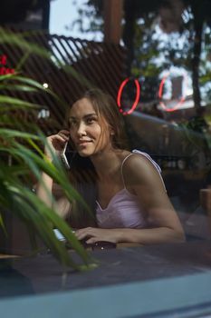 young asian pretty woman sitting at table of coffee shop, calling by smartphone. beautiful smiling lady using cell phone shot through window glass outside of cafe. modern communication technology