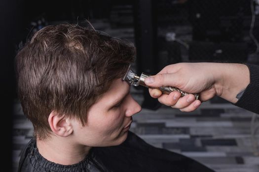 The hand of a barber or hairdresser with a portable clipper cuts the client's man with dark long hair. Hairdressing.